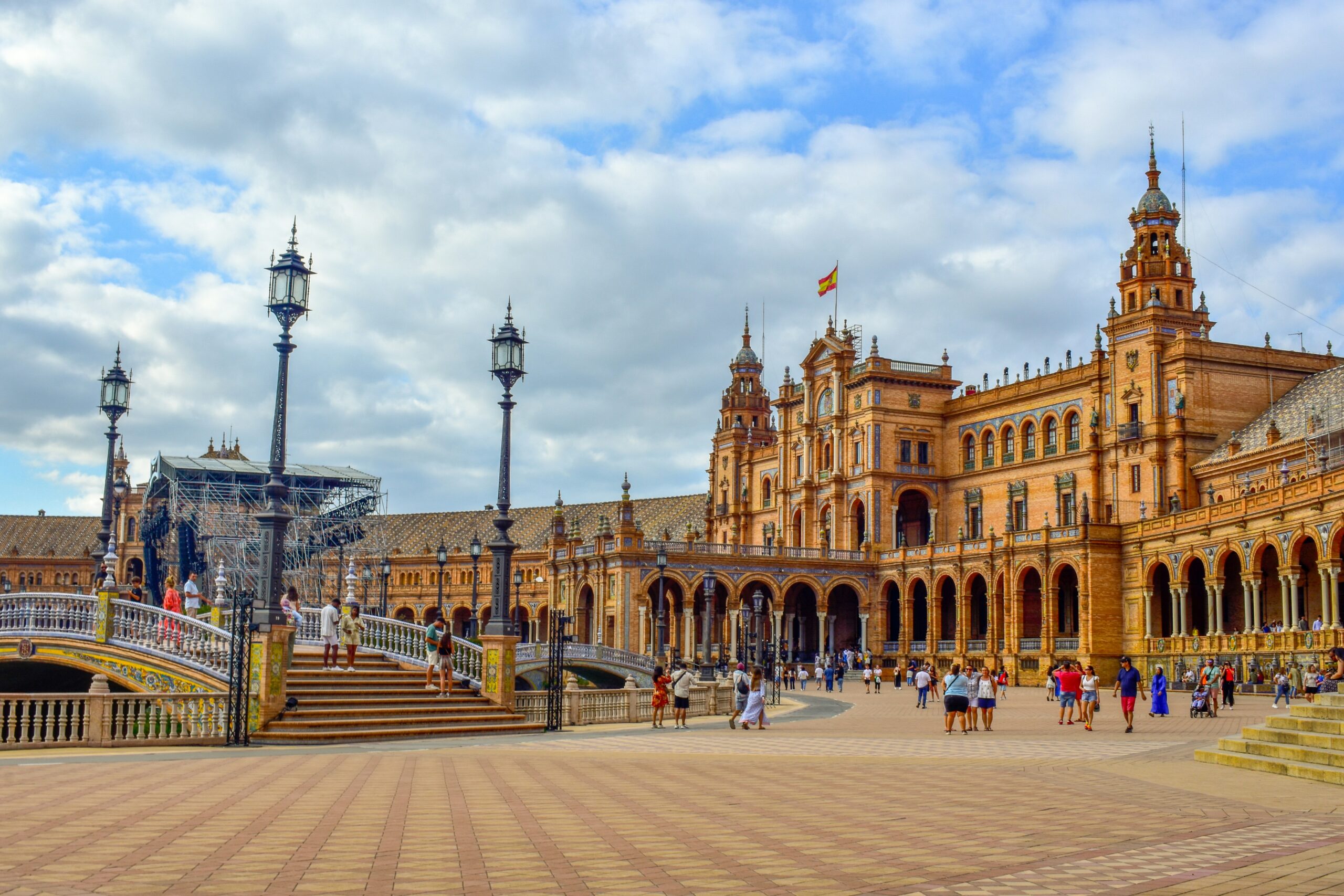 Wide-angle view of the ornate Neo-Mudéjar architecture of Plaza de España in Seville, Spain, featuring a brick courtyard, bridges with blue-and-white tiling, and a partly cloudy sky.