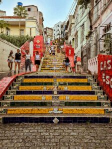 A perspective view of the Escadaria Selarón in Rio de Janeiro, Brazil, showing the famous staircase covered in vibrant yellow, green, and blue mosaic tiles with several tourists and locals on the steps.