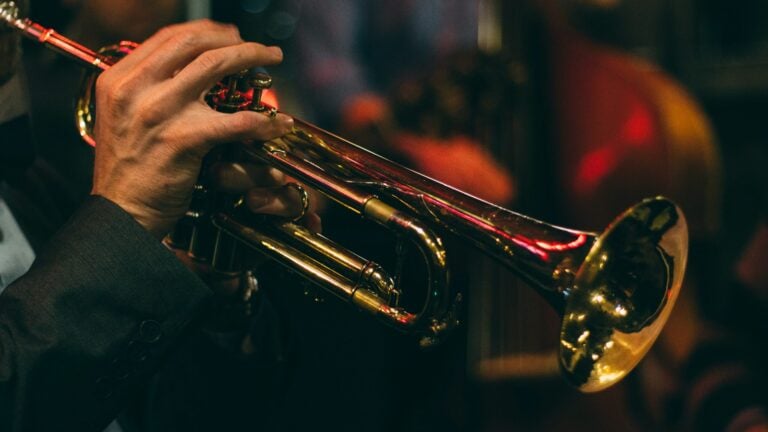 A close-up, atmospheric shot of a musician’s hands playing a brass trumpet. The gold of the instrument reflects warm red and orange stage lights, with the valves and tubing in sharp focus against a dark, blurred background of a live jazz ensemble.