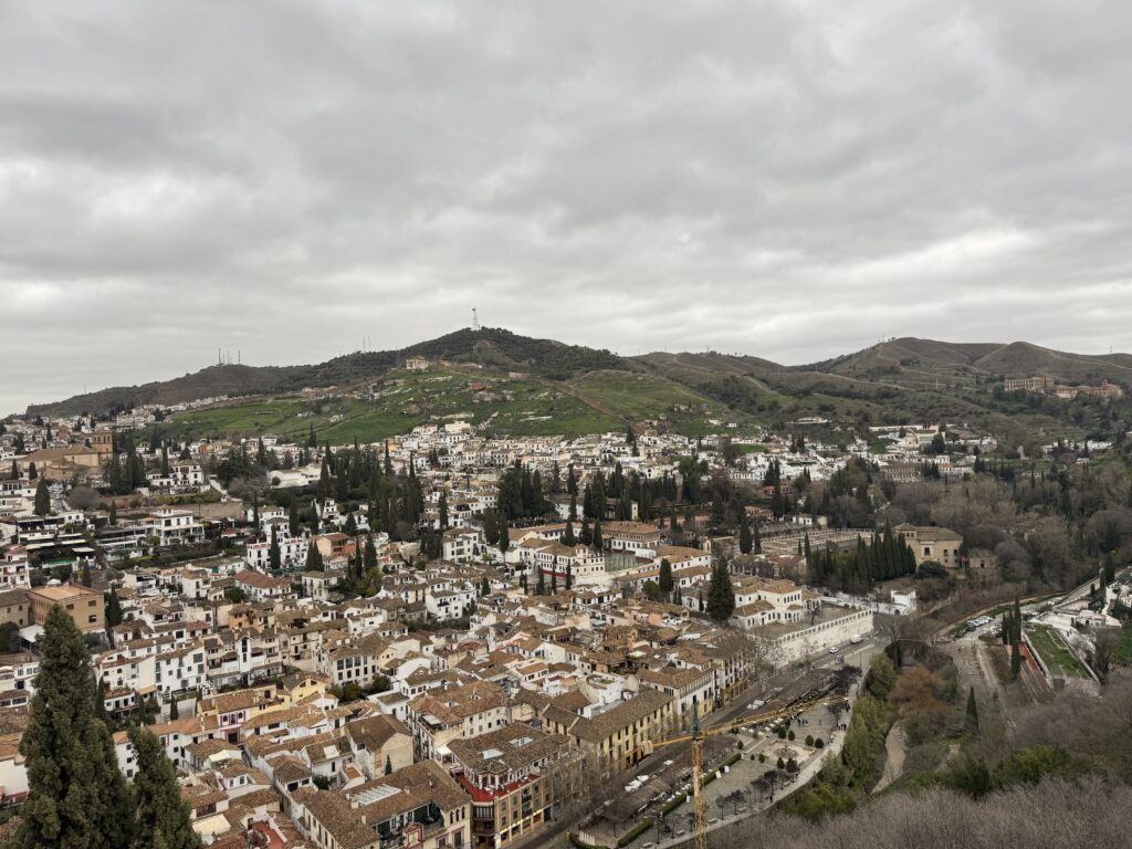 High angle view over the manicured hedge mazes of the Generalife gardens looking out toward the historic fortress walls under a cloudy sky.