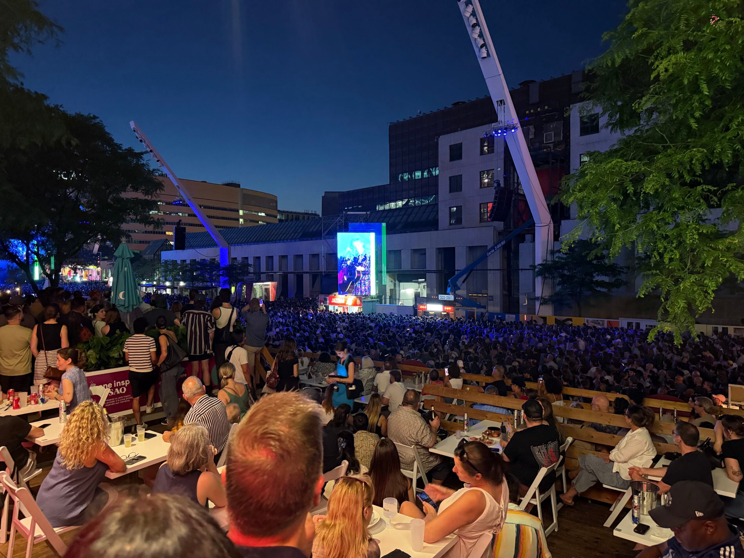 A nighttime audience fills the outdoor Montreal Jazz Festival site, with an illuminated concert screen and downtown buildings