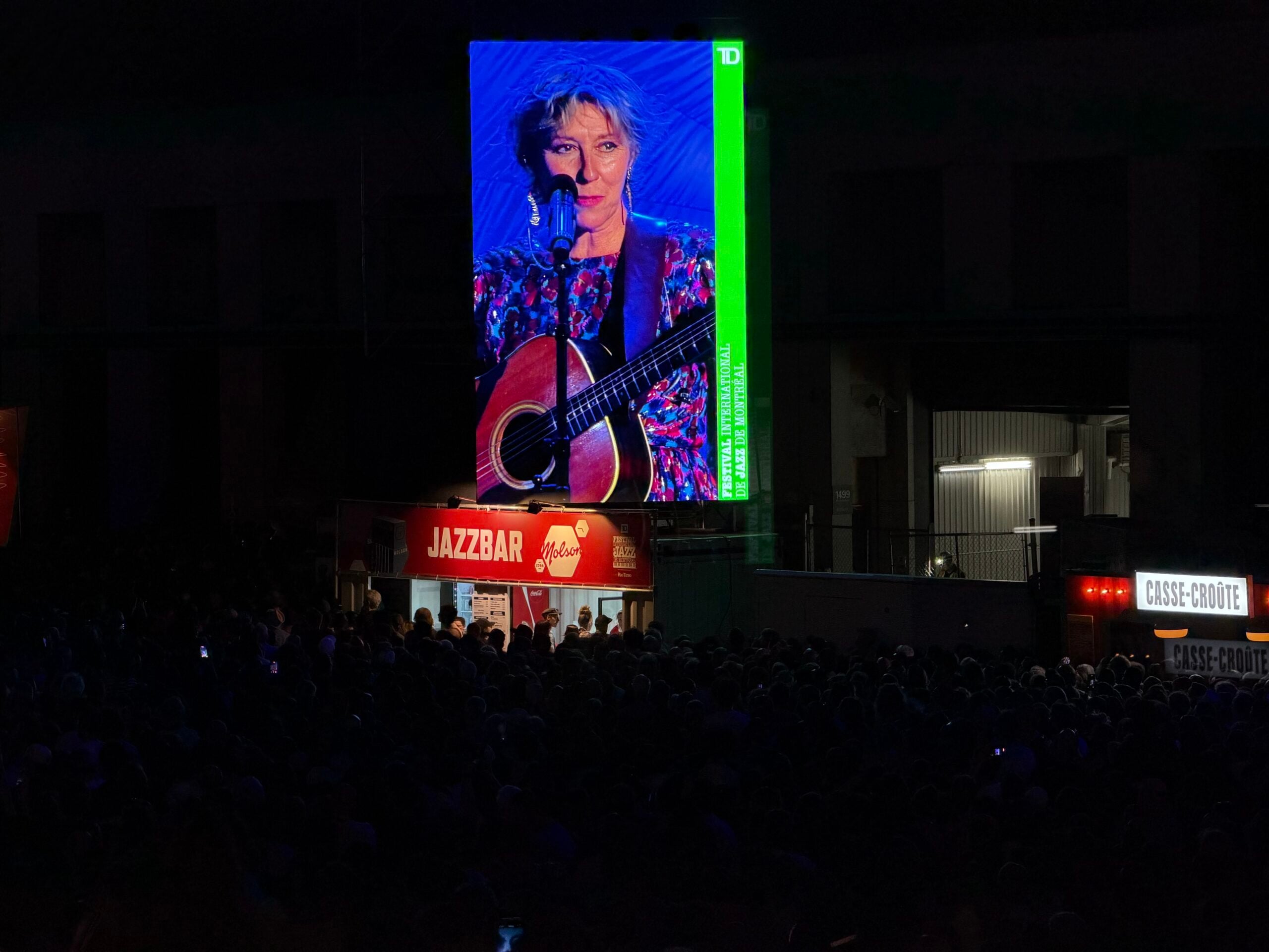 A large festival screen shows a singer playing guitar above a nighttime outdoor audience at the 2026 Montreal Jazz Festival